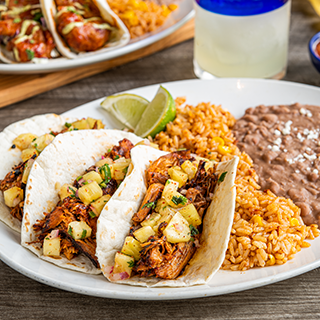 Tacos Al Pastor: Seasoned, braised carnitas and pineapple salsa in hand-pressed flour tortillas. Served with Mexican rice and choice of beans.